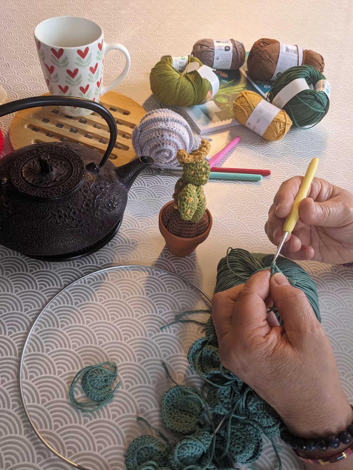 Une main crochète une petite pièce de laine verte, à côté d'une théière et de pelotes